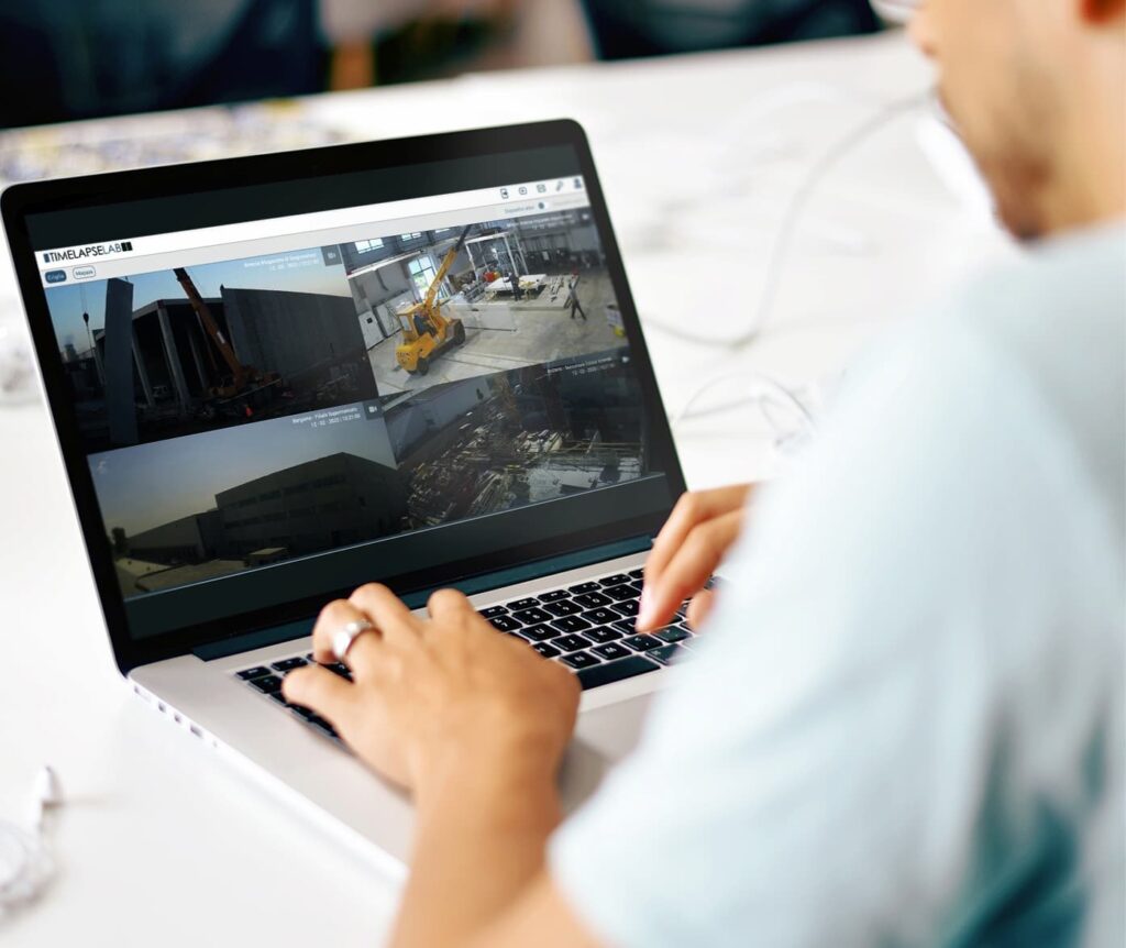 Una persona di cui si vedono bene solo le mani è seduta alla scrivania con davanti un portatile Macbook. Sullo schermo sono visualizzate quattro schermate diverse che rappresentano immagini di quattro diversi cantieri